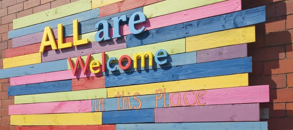 A colourful sign of wooden boards and 3D printed letters which reads "All are welcome in this place"