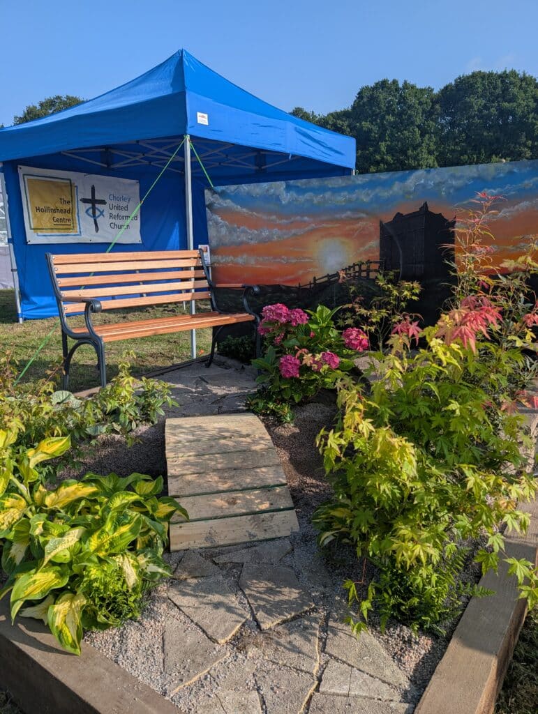 A picture of a small show garden at Chorley flower show. The garden has a low arch bridge over a small stream leading to a park bench with Japanese style planting around it. In the background is a gazebo with logos of Chorley URC and the Hollinshead Centre